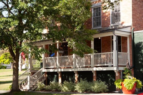 A brick building with a porch, surrounded by trees and greenery, featuring a welcoming entrance and outdoor seating.