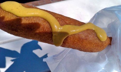 A close-up of a corn dog drizzled with mustard, held in a hand with a shadow visible on the surface.