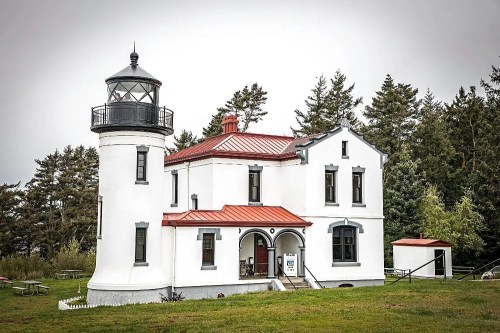 A white lighthouse with a red roof, surrounded by trees and grass, featuring a lantern room and a welcoming entrance.