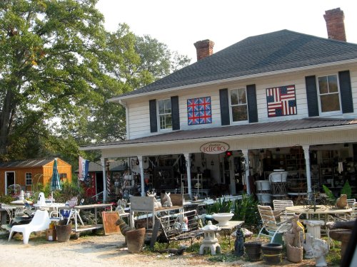 A quaint shop with colorful quilt patterns on the windows, surrounded by various outdoor items and greenery.