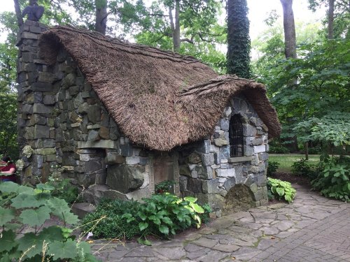 A quaint stone cottage with a thatched roof, surrounded by lush greenery and trees.