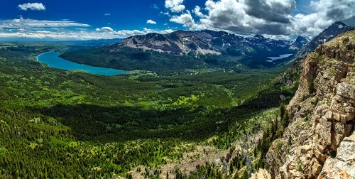 A panoramic view of lush green valleys and mountains under a blue sky with scattered clouds, featuring a winding lake.