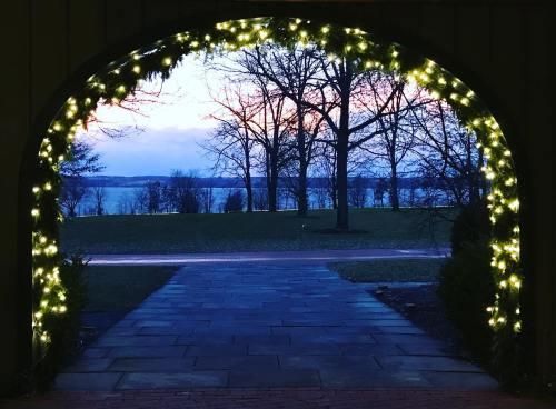 A view through an archway adorned with lights, overlooking a lake and trees at dusk.