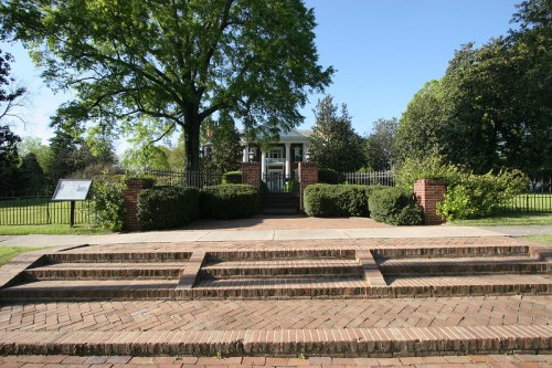 A brick pathway leads to a historic house surrounded by greenery and trees, with a fence in the background.