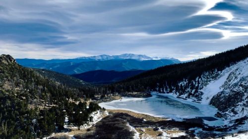 A serene winter landscape featuring a frozen lake surrounded by snow-covered mountains and pine trees under a cloudy sky.