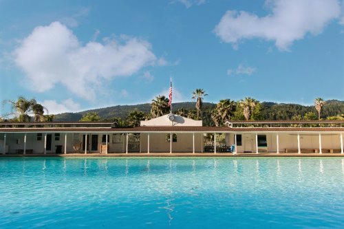 A serene pool scene with a building in the background, surrounded by palm trees and mountains under a clear blue sky.