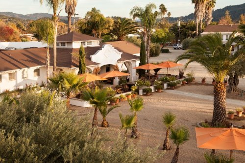 A scenic view of a courtyard with palm trees and orange umbrellas outside a charming building in a sunny landscape.