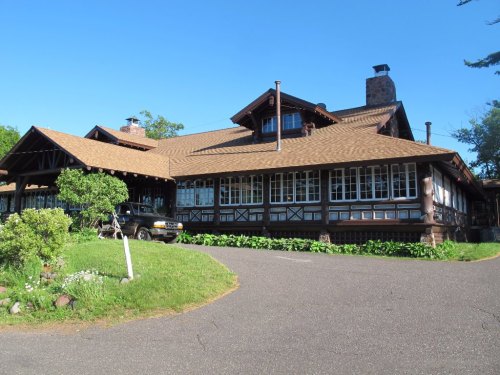 A large, rustic wooden house with a sloped roof, surrounded by greenery and a paved driveway.