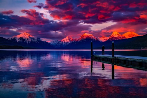 A person stands on a dock at sunset, with vibrant clouds reflecting on a calm lake and snow-capped mountains in the background.
