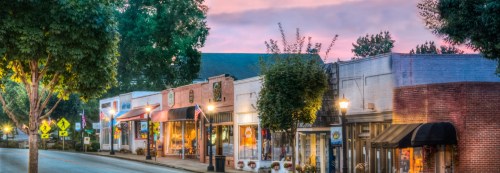 Charming small-town street at dusk, featuring shops, trees, and a colorful sky.