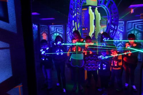 A group of children in a neon-lit laser tag arena, holding laser guns and surrounded by colorful lights.