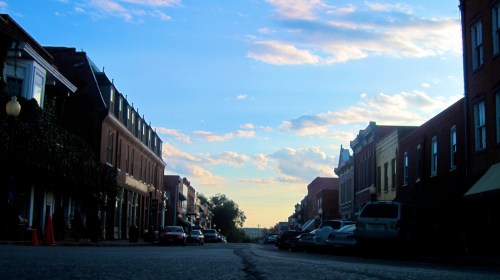 A quiet street lined with historic buildings, under a blue sky with scattered clouds at sunset.