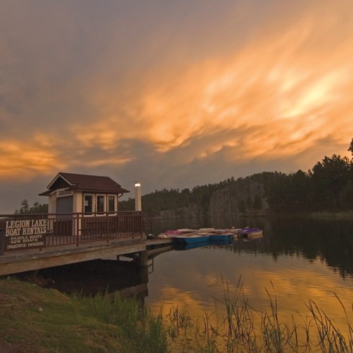A serene lake scene featuring a boat rental shack, calm waters, and a cloudy sky at dusk.