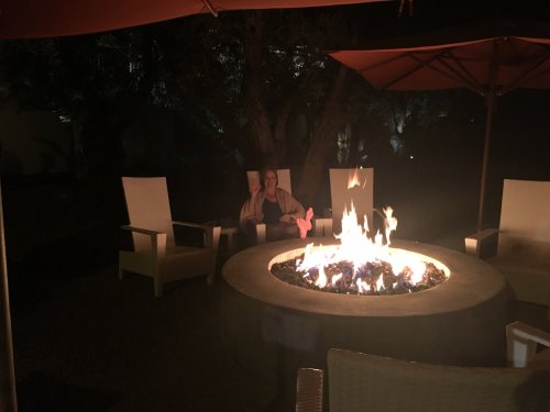 A cozy outdoor setting with a fire pit surrounded by chairs, and a person sitting nearby in the dim light.
