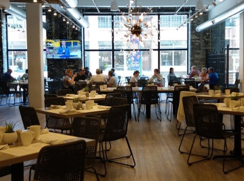 A modern restaurant interior with tables set for dining, patrons enjoying meals, and a large window with a view outside.