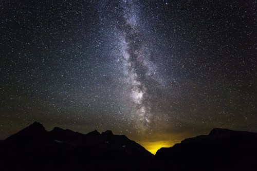 A stunning night sky filled with stars and the Milky Way, silhouetted against dark mountains and a glowing horizon.