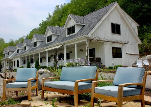 A white cottage with multiple gabled roofs, surrounded by greenery, features blue chairs in the foreground.