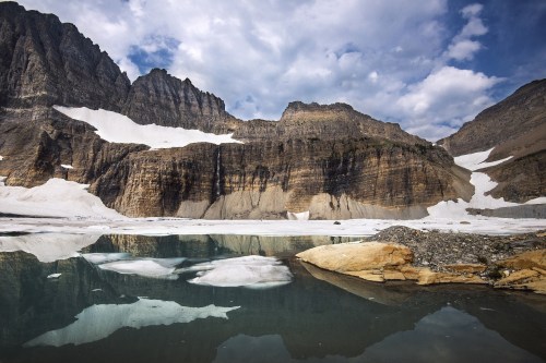 A serene mountain landscape with rocky cliffs, snow patches, and a calm lake reflecting the sky.