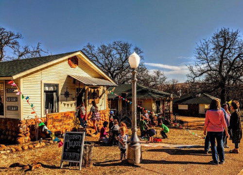 A group of children and adults gather outside a charming house decorated with colorful bunting and a sign for a toy store.