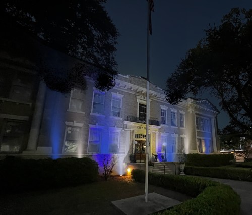 Historic city building illuminated in blue at night, with a flagpole in front and lush greenery surrounding it.