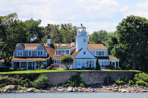 A charming lighthouse beside a large coastal house, surrounded by greenery and water under a clear blue sky.