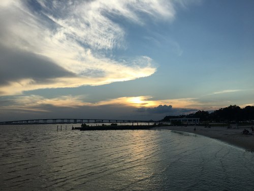 Sunset over a calm beach with a bridge in the distance and clouds in the sky, creating a serene atmosphere.