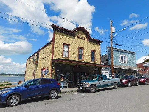 A quaint, two-story building with a shop on the ground floor, surrounded by parked cars and a scenic view of the water.
