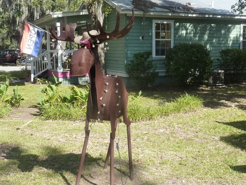 A metal reindeer sculpture adorned with lights stands in a grassy yard in front of a green house.