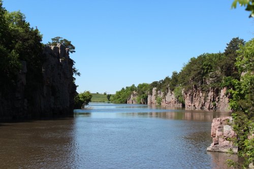 A serene river flows between rocky cliffs, surrounded by lush greenery under a clear blue sky.