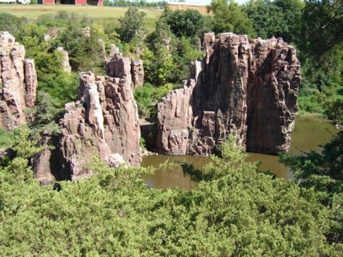 Rock formations rise from a serene pond, surrounded by lush greenery and trees under a clear blue sky.