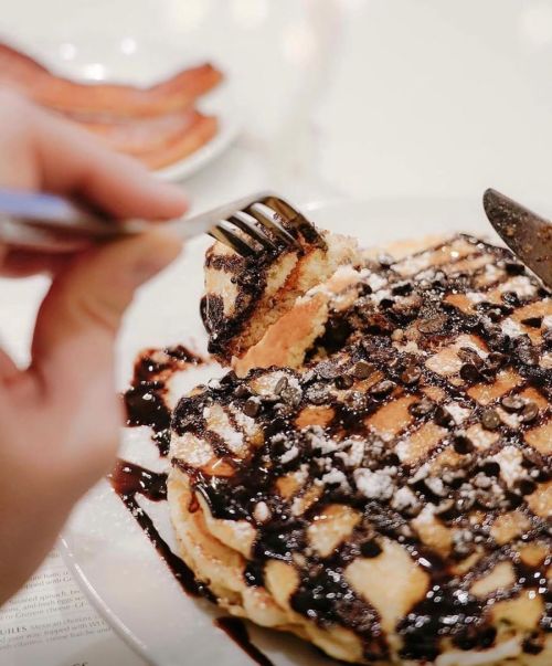 A hand holding a fork lifts a piece of chocolate-drizzled pancakes topped with chocolate chips and powdered sugar.