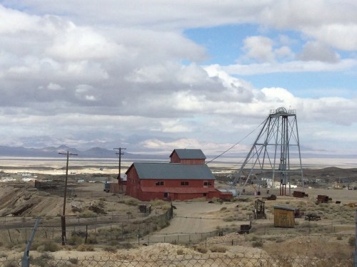A red mining building with a tall structure nearby, set against a cloudy sky and distant mountains.
