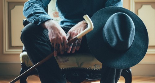 A person sits on a chair, holding a cane, with a wide-brimmed hat resting beside them.