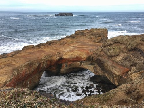 A rocky coastal landscape featuring an arch formation with waves crashing below and a distant island in the background.