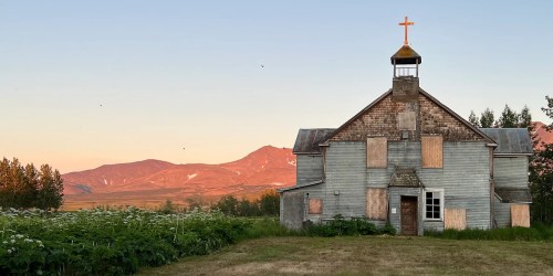 Abandoned church with a cross, surrounded by greenery and mountains at sunset.