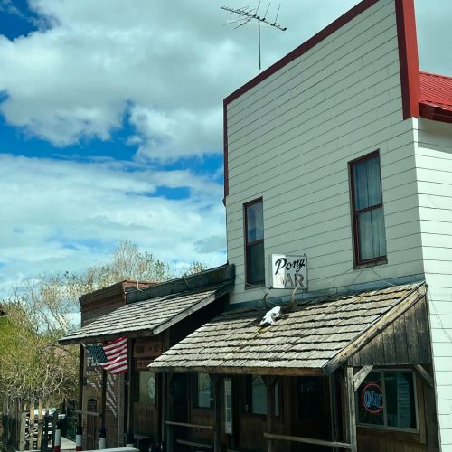 A rustic bar with a wooden facade, sign reading "Pony Bar," and an American flag outside under a partly cloudy sky.