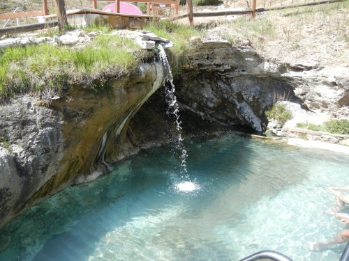 A serene natural pool with a small waterfall cascading into clear water, surrounded by rocky terrain and greenery.