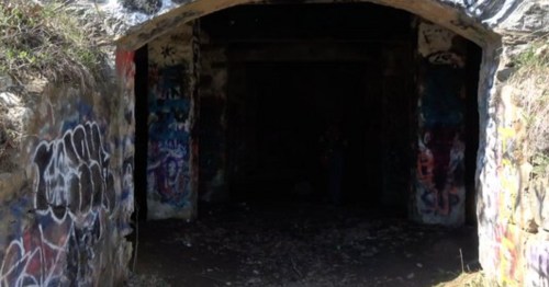 A dark tunnel entrance surrounded by colorful graffiti on stone walls, with overgrown grass at the edges.