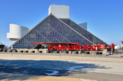 Exterior view of the Rock and Roll Hall of Fame, featuring a large "LONG LIVE ROCK" sign in front. Clear blue sky above.