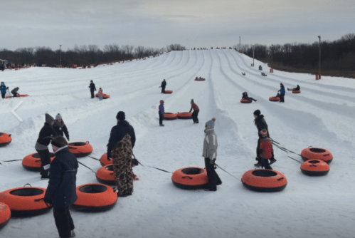 People enjoying a snow tubing hill, with orange tubes scattered on the snowy slopes under a cloudy sky.