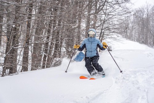 A skier navigates through deep snow in a forested area, wearing a blue jacket and orange skis. Snowflakes are falling.