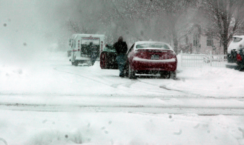 A person struggles to enter a car in a snowy, blizzard-like scene with heavy snowfall and limited visibility.