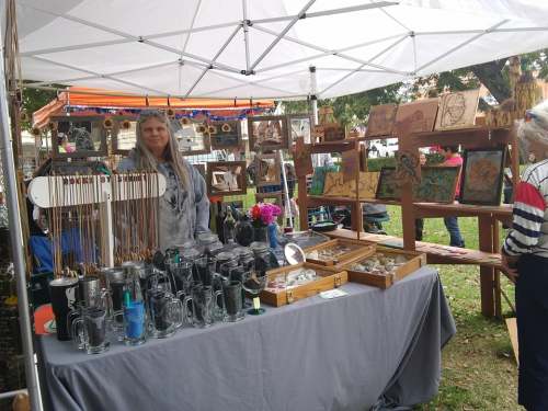 A vendor at a market displays handmade jewelry and crafts under a tent, with customers browsing nearby.