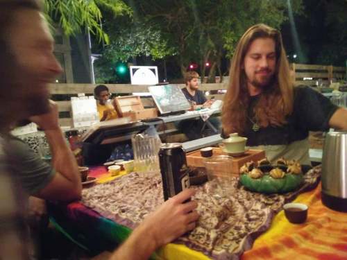 A group of people socializing at a table with drinks and snacks, surrounded by greenery and a relaxed atmosphere.