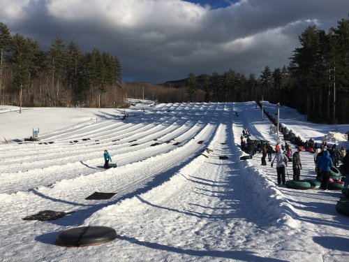 A snowy tubing hill with multiple lanes, people sliding down, and trees in the background under a cloudy sky.