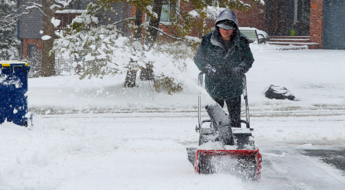 A person using a snowblower to clear a snowy driveway on a winter day. Snow is blowing in the air around them.