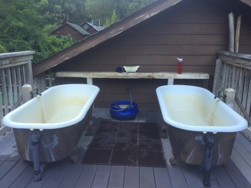 Two vintage bathtubs on a wooden deck, with a bench and a blue bucket nearby, surrounded by greenery.