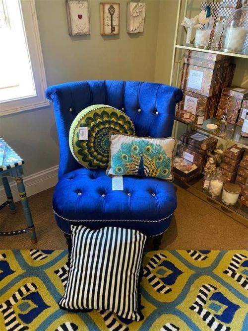 A vibrant blue tufted chair with decorative pillows, set against a colorful patterned rug and a shelf of gift items.