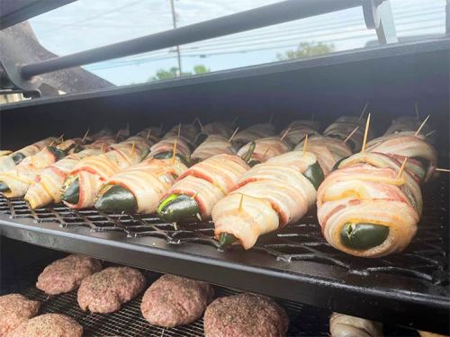Bacon-wrapped jalapeños on a grill, with burgers cooking below them.