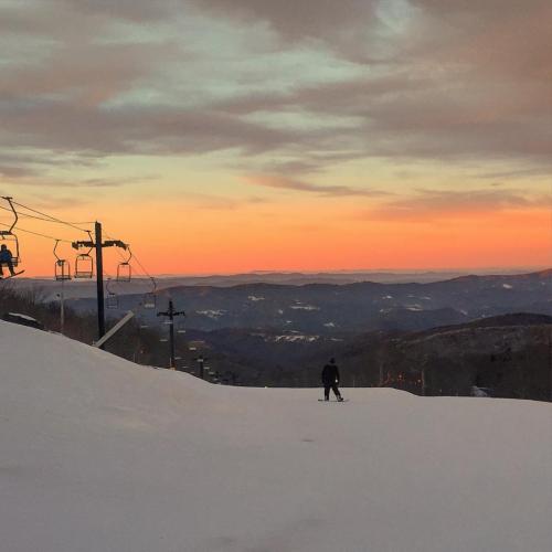 A skier on a snowy slope at sunset, with ski lifts in the background and mountains fading into the horizon.